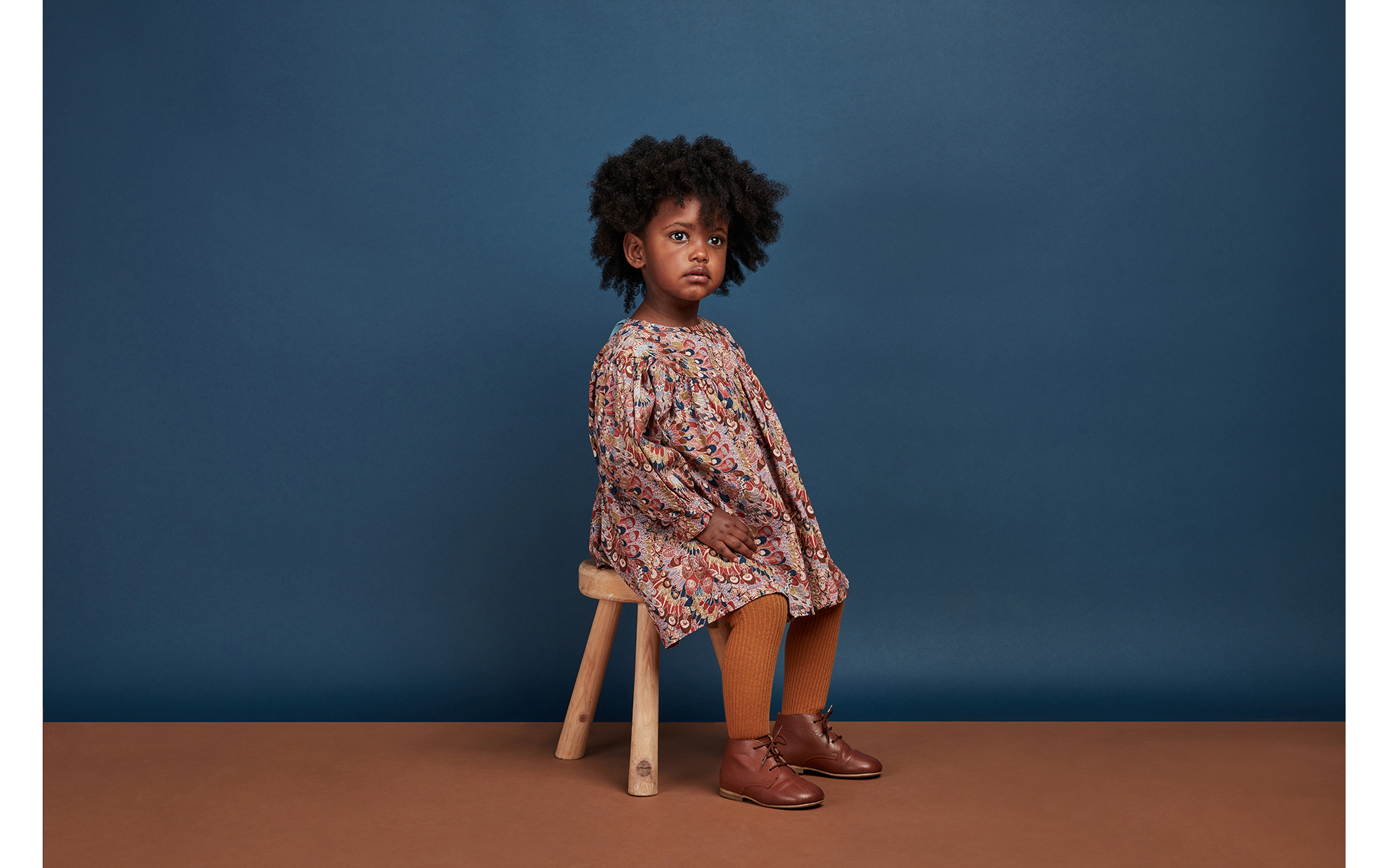 child girl model sitting on wooden stool looking away in front of Oxford blue back drop brand children photo shoot