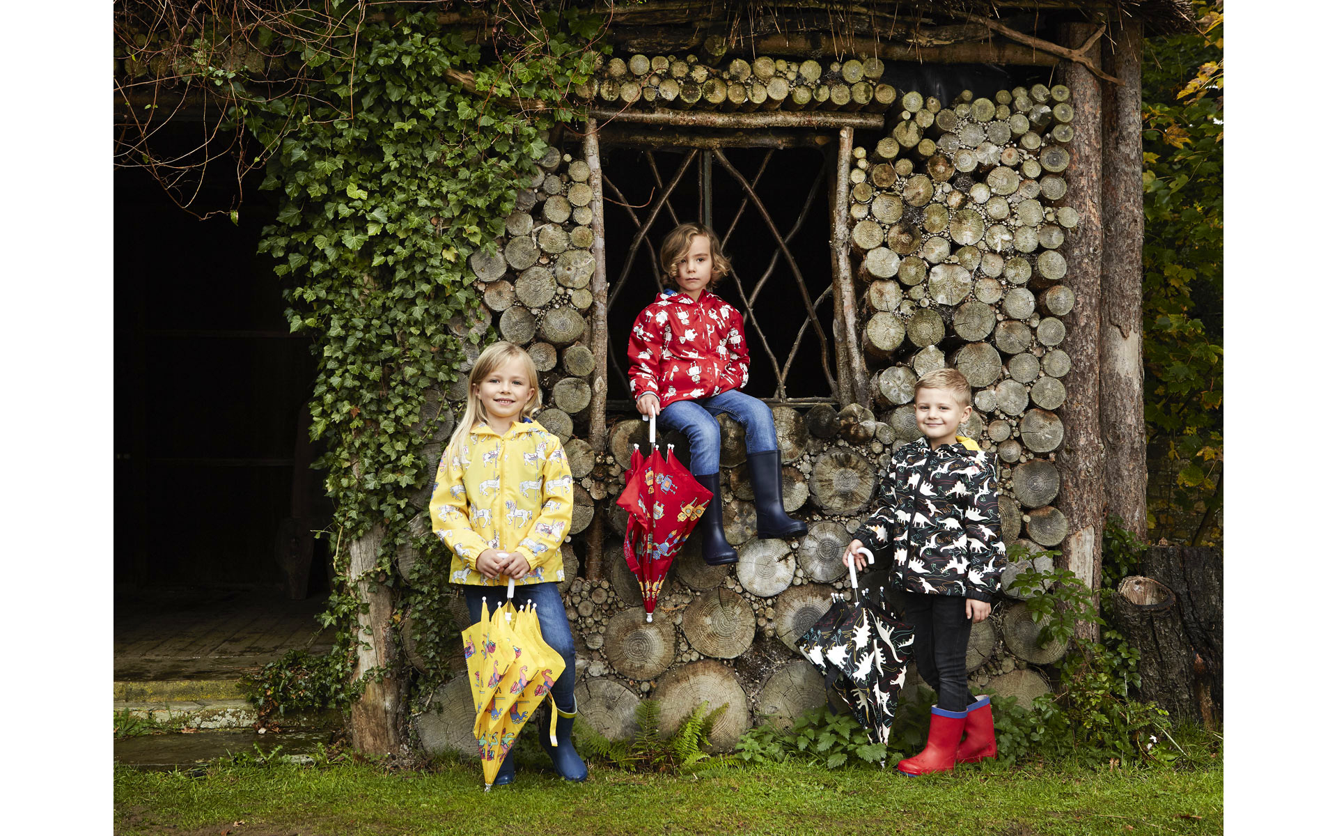 3 child models in rain coats next to wooden house in Norfolk country in winter on a brand fashion photo shoot